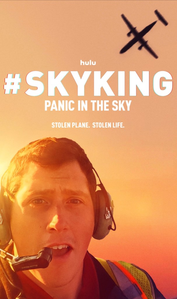 A man wearing a headset and safety vest speaks into a microphone, with an airplane flying in the sky above him. The backdrop features a sunset.